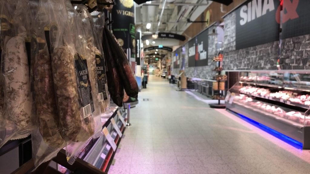 An empty aisle in a brightly lit Finnish supermarket, with shelves full of sausages and groceries but no customers or staff in sight.