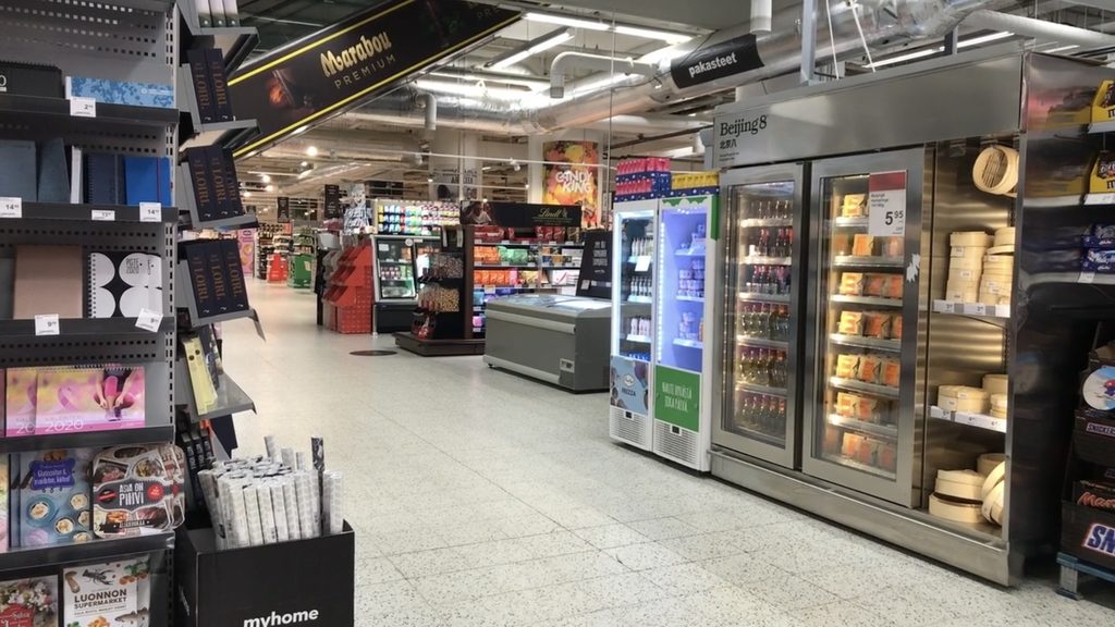 An empty aisle in a brightly lit Finnish supermarket, with shelves full of books and beverages and groceries but no customers or staff in sight.