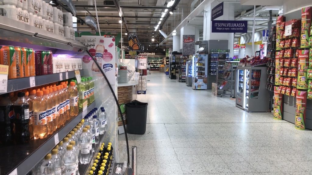 An empty aisle in a brightly lit Finnish supermarket, with shelves full of drinks and groceries but no customers or staff in sight.